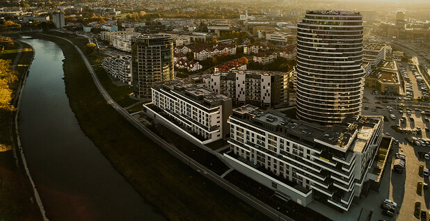 Zdjęcie lotnicze nowoczesnego kompleksu mieszkalno-biurowego Capital Towers w Rzeszowie usytuowanego nad brzegiem rzeki Wisłok. Po prawej stronie dominuje wysoki, charakterystyczny wieżowiec o zaokrąglonej bryle, obok którego stoją niższe, kaskadowe budynki wielorodzinne w czarno-białej kolorystyce. W tle widoczna jest rozległa panorama miasta Rzeszów, parkingi samochodowe oraz otaczająca rzekę zieleń w świetle zachodzącego słońca.